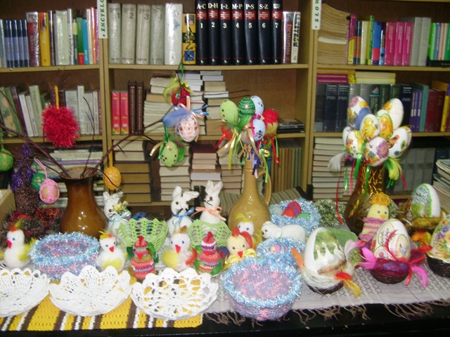 Zdjęcie przedstawiające ozdoby i dekoracje wielkanocne m.in. pisanki, kurczaczki i koszyczki. W tle regały z książkami.