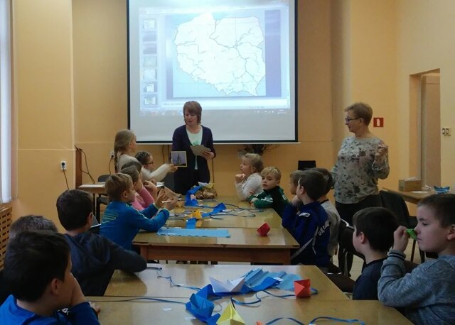 Uczniowie siedzą przy stolikach, na których leżą wykonane z papieru prace. W tle wyświetlona mapa Polski.