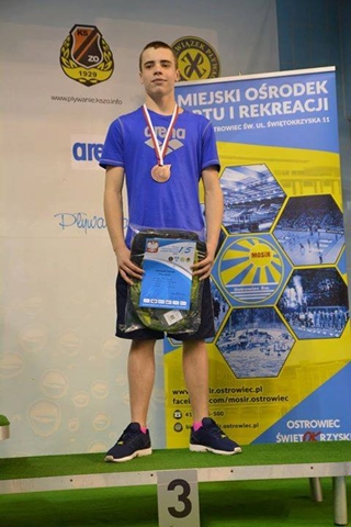Zdjęcie przedstawia zdobywcę trzeciego miejsca w Mistrzostwach Polski w Pływaniu. Chłopiec stoi na podium i prezentuje medal i nagrodę rzeczową.