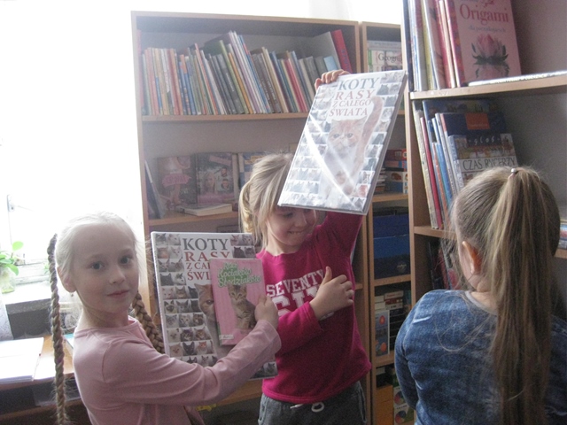 Dzieci prezentują książki o kotach. W tle regały z książkami.