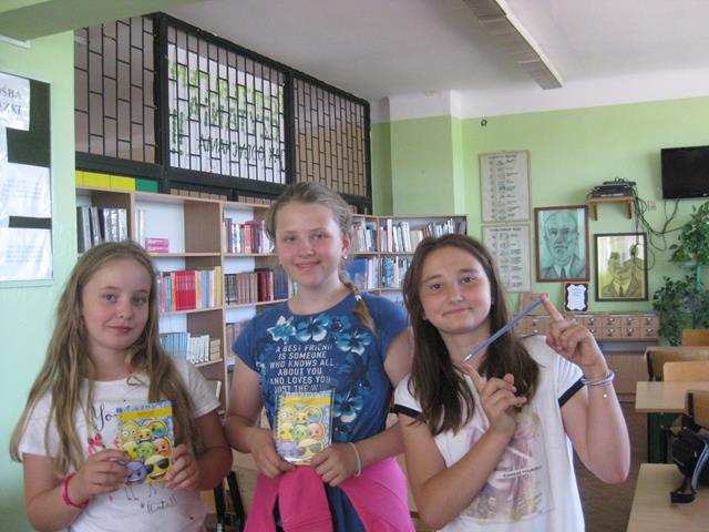 Trzy uczennice w bibliotece szkolnej. Dwie dziewczynki trzymają w dłoniach zdjęcia kolorowych emotikonów, a trzecia prezentuje ołówek.