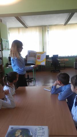 Zdjęcie przedstawia nauczycielkę i dzieci podczas zajęć w bibliotece szkolnej. Kobieta prezentuje uczniom treści z otwartej książki.
