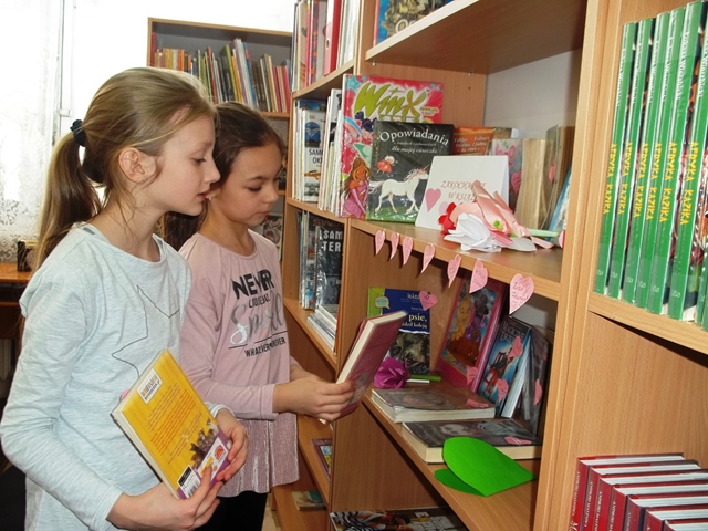 Dwie uczennice wybierają książki z półki regału bibliotecznego.