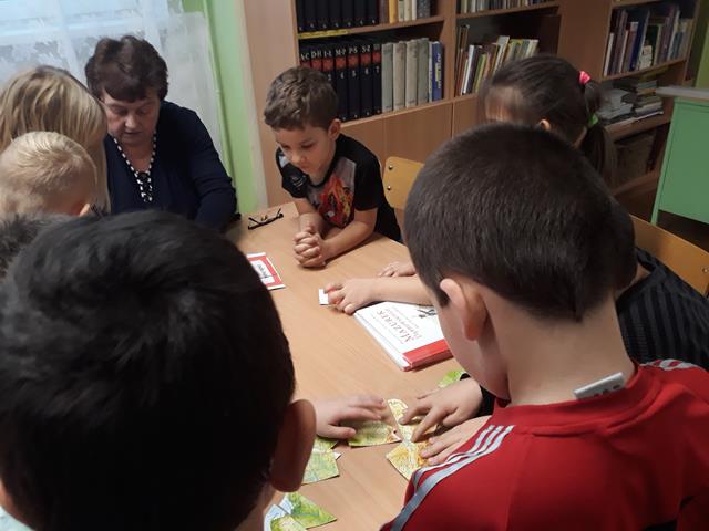 Grupa uczniów z panią ze świetlicy siedzą przy stoliku. Kilku chłopców układa puzzle mapy Polski.
