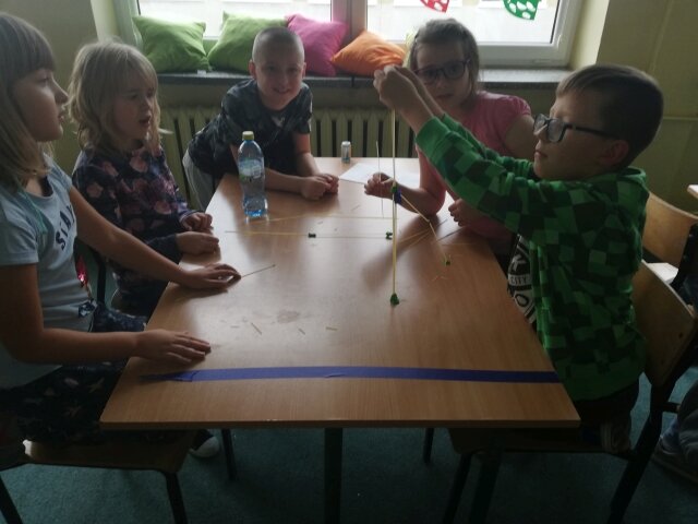 Pięcioro uczniów siedzi przy stoliku. Jeden z nich wykonuje doświadczenie, które pozostałe dzieci obserwują.