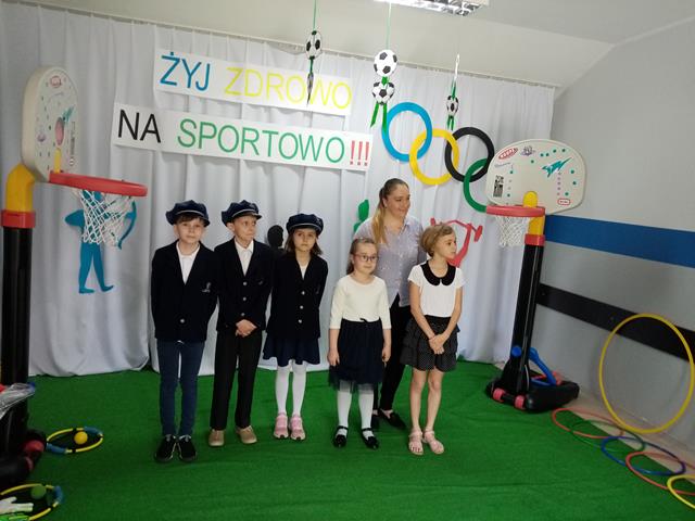 Pięcioro dzieci w strojach galowych z panią opiekunką. W tle dekoracja o tematyce sportowej z hasłem „Żyj zdrowo na sportowo!!!”