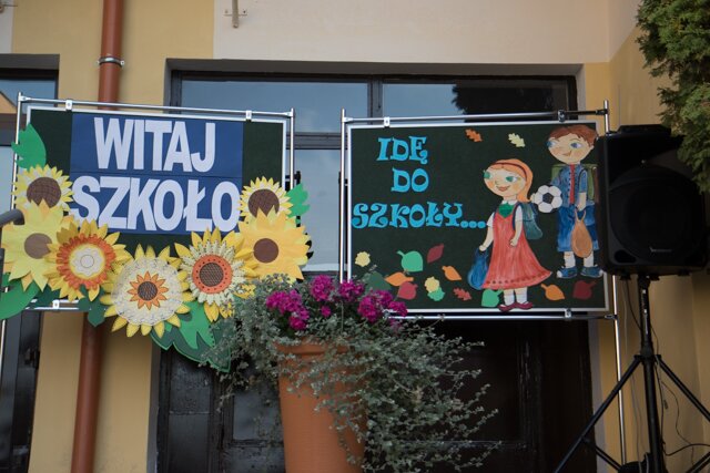 Zdjęcie przedstawia dwie tablice na tle wejścia do szkoły. Na jednej z ich napis: ,,Witaj szkoło’’ oraz wycięte z papieru słoneczniki. Na drugiej tablicy widnieją wycięte postaci chłopca i dziewczynki idących do szkoły i napis: ,,Idę do szkoły’’.