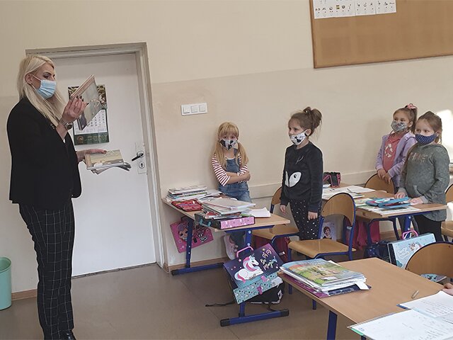 Pani dyrektor eksponuje książki uczniom, którzy stoją przy swoich ławkach.