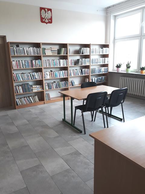 W szkolnej bibliotece na środku stoi stół, przy nim trzy czarne krzesła. Przy ścianie znajduje się regał z książkami, a nad nim wisi godło Polski. Po prawej stronie jest okno i parapet, na którym stoją kwiatki.
