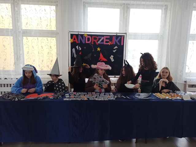 Zdjęcie przedstawia uczniów klas starszych w andrzejkowych przebraniach. Na stole przykrytym granatowym suknem leżą porozkładane akcesoria do wróżb. W tle tablica z napisem: ,,Andrzejki”.