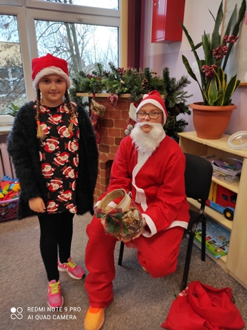Zdjęcie przedstawia uczennicę w mikołajowej czapce. Dziewczynka pozuje do zdjęcia z Mikołajem. W tle świątecznie przystrojony kominek.