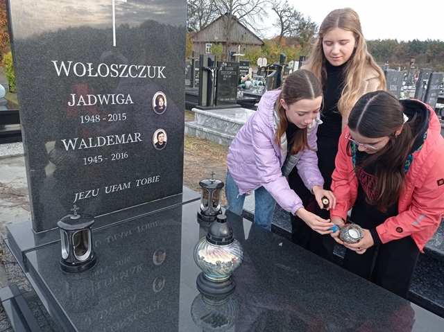 Zdjęcie przedstawia trzy uczennice zapalające znicz. Dzieci odwiedzają groby zmarłych nauczycieli byłych pracowników Szkoły Podstawowej nr 8 im. Bolesława Zygmunta Wirskiego w Chełmie.