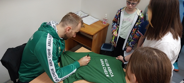 Zdjęcie przedstawia zawodnika klubu “Arka Chełm”, który podpisuje uczennicom klubową koszulkę w kolorze zielonym.