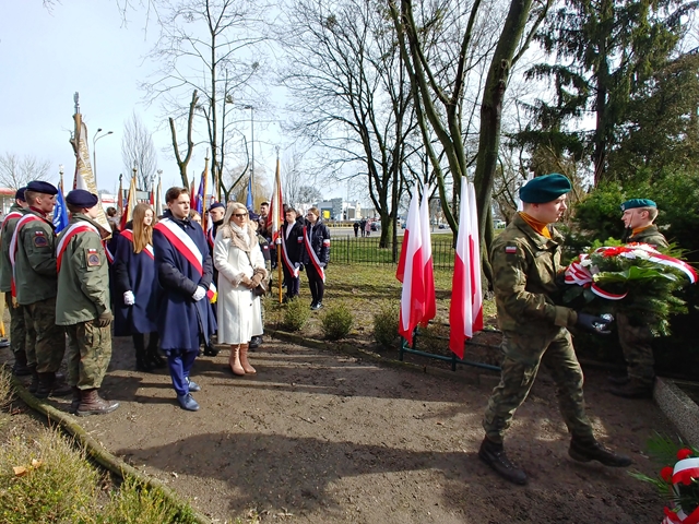 Zdjęcie przedstawia delegacje żołnierzy, nauczycieli i uczniów podczas uroczystości na cmentarzu. Żołnierze składają wieniec. W tle biało-czerwone flagi.