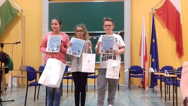 Zdjęcie przedstawiające uczniów prezentujących nagrody i dyplomy za udział w konkursie. W tle flaga Polski i Unii Europejskiej.