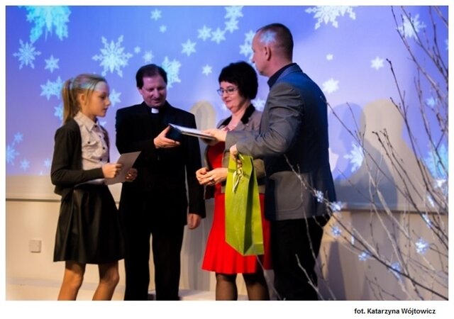 Zdjęcie przedstawia moment wręczania naszej laureatce dyplomu za udział w Chełmskim Konkursie Kolęd i Pastorałek. Na zdjęciu znajduje się uczennica oraz inne osoby wręczające nagrody, w tle widoczne spadające śnieżynki.