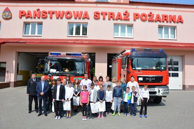 Zdjęcie przedstawia grupę dorosłych i dzieci pozujących przed budynkiem Państwowej Straży Pożarnej. W tle wozy strażackie.